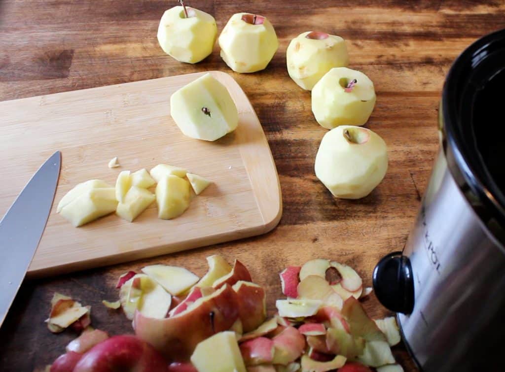 freshly peeled apples are being chopped up and placed into a crockpot. there is a mess of peels across the wooden table top!