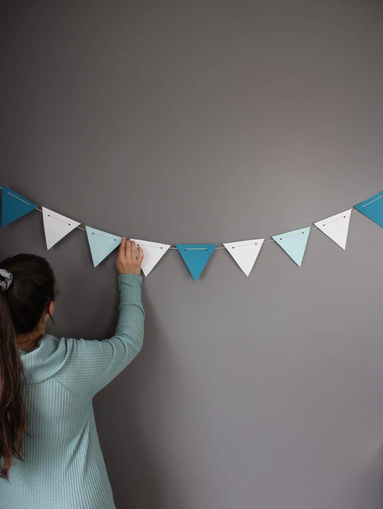 a woman in a light blue sweater with a pony tail held up by a grey scrunchie hangs party decor made of dark blue, light blue, and white card stock.