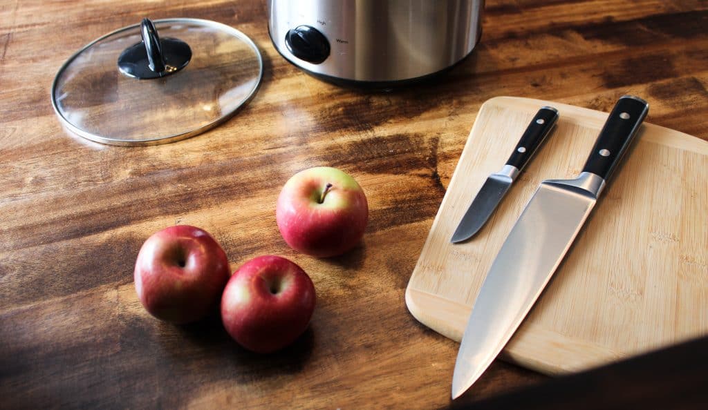 a crockpot, knives, and a cutting board wait next to some red apples to begin making crockpot applesauce!