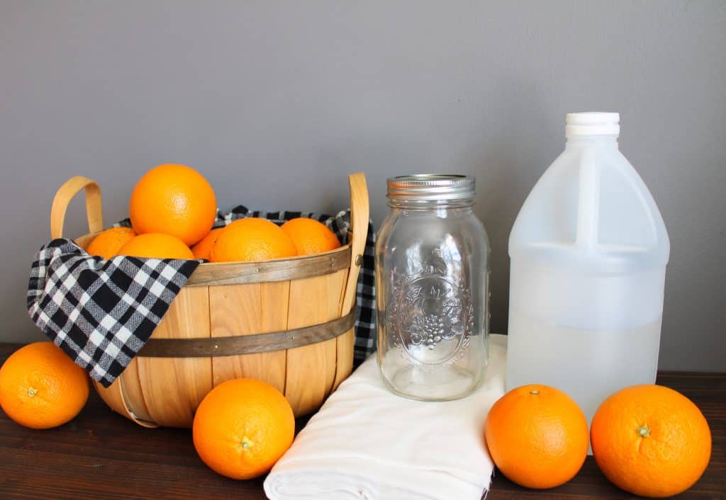 a wooden basket lined with a black, grey, and white plaid tea towel overflows with oranges next to a large mason jar, cheesecloth, and a jug of vinegar. oranges are scattered across a wooden countertop