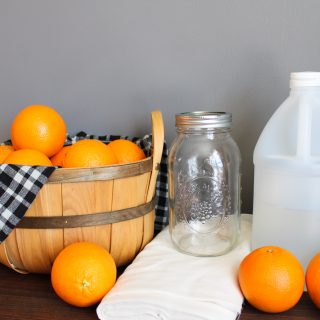 a wooden basket lined with a black, grey, and white plaid tea towel overflows with oranges next to a large mason jar, cheesecloth, and a jug of vinegar. oranges are scattered across a wooden countertop