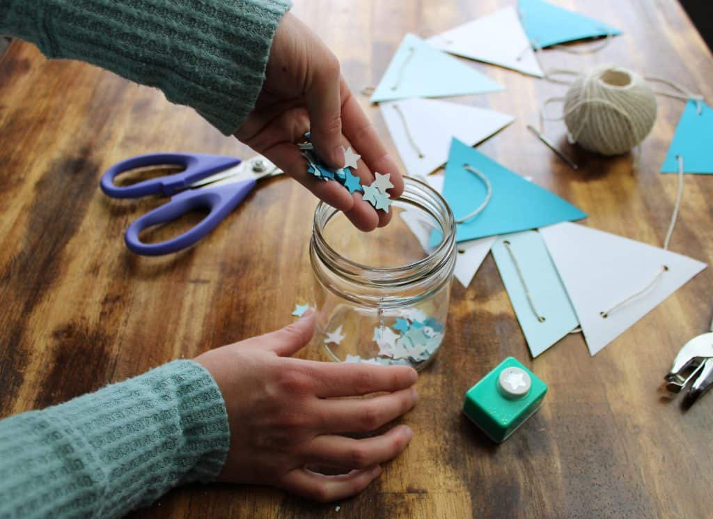 a woman stores the little stars she punched out of the scrap paper from her project in a glass jar. the table is covered with the tools she used for her project and a completed paper bunting.