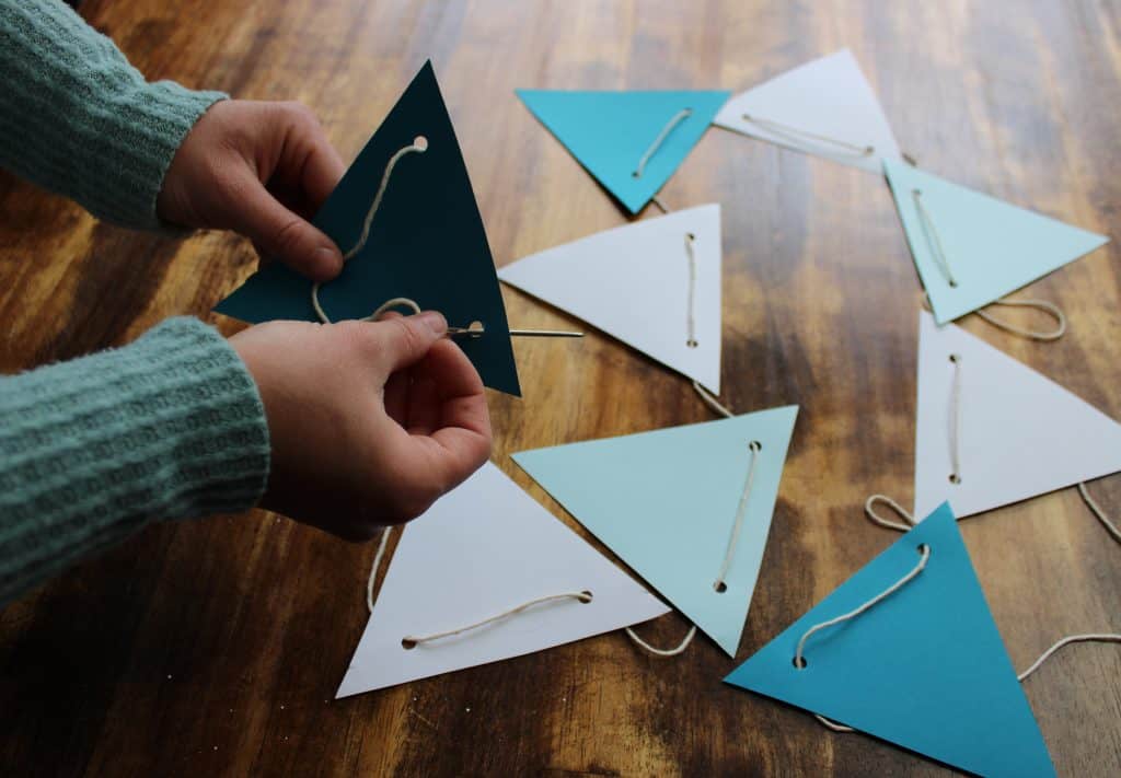 a woman uses a large needle to thread string through the hole punches in each triangle. she is stringing the bunting together!