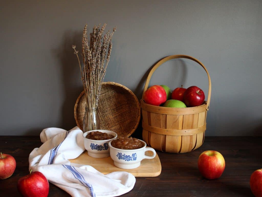 applesauce is displayed in lovely antique cups with dried lavender and a bushel of apples behind it. they sit on top of a cutting board with a tea towel next to it and a few apples scattered about.