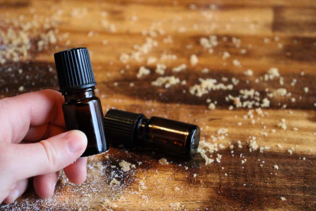 a woman holds a bottle of essential oils while another bottle is laying on the table behind it. there is sugar scattered across the wooden table top.