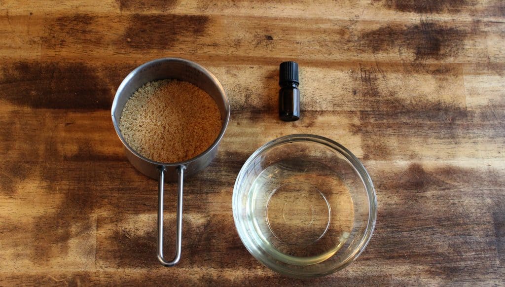 a metal measuring cup filled with brown sugar, a bottle of essential oil, and melted coconut oil in a glass bowl.