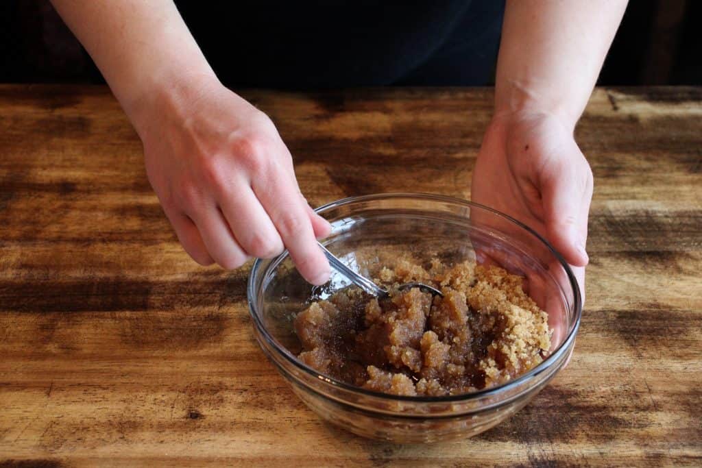 the woman uses a silver spoon to mix the brown sugar and coconut oil together