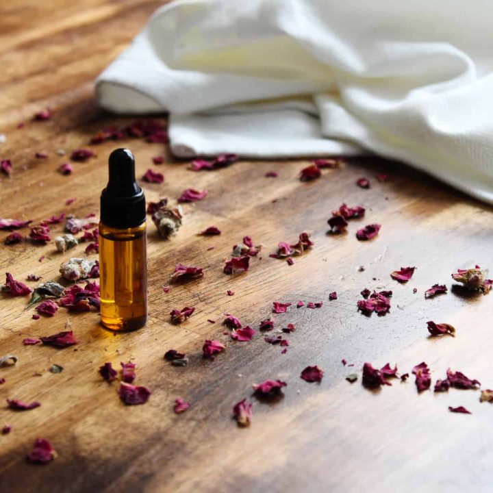 an amber glass dropper bottle with rose petals surrounding it and a white fabric cloth nearby