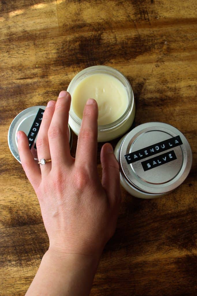 a woman's hand with a wedding ring on reaching for some calendula salve. there are two labelled jars. one is open
