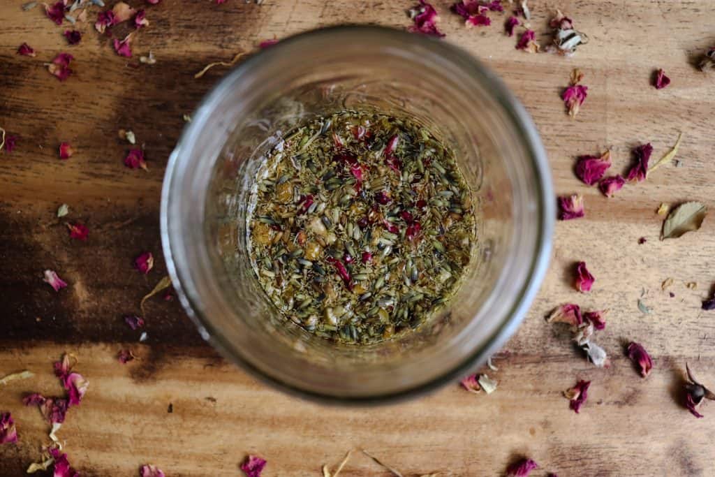 a photo from above of herbs infusing in oil in a glass jar. rose petals are all around.
