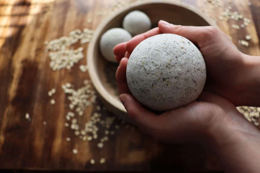 a woman holds a large bath bomb in her hands in front of the others that are in a wooden bowl. there are oats and dried lavender scattered about.