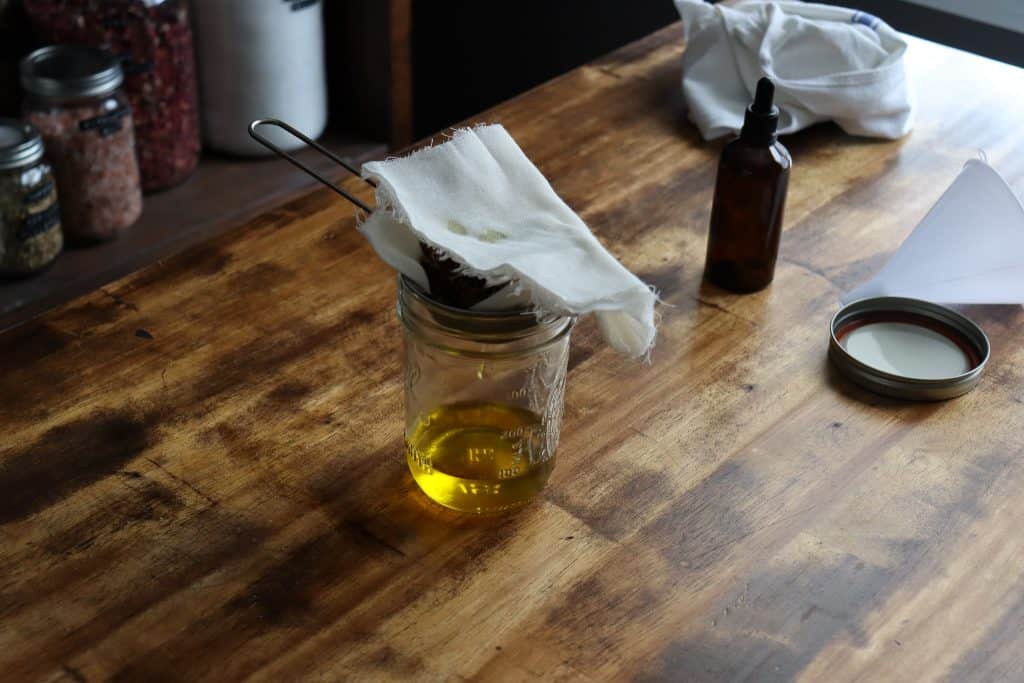 herbs are in cheesecloth sitting inside a mesh strainer over a glass jar. the oil is dripping out.