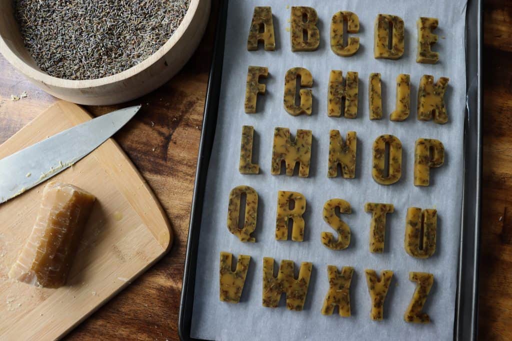the letters are set and out of the mold on a parchment lined tray. to their left there is a block of beeswax on a cutting board with a knife and a wooden bowl with dried lavender in it.