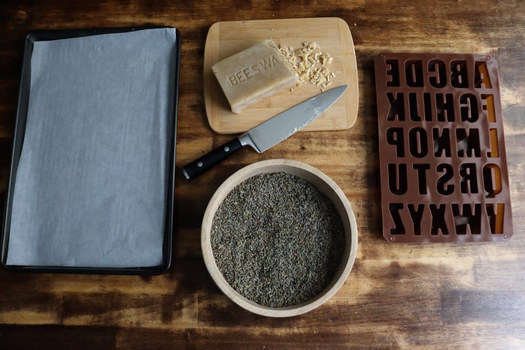 from left to right: a baking sheet with parchment paper cut to size in it, a wooden cutting board with beeswax and a knife on it, dried lavender in a wooden bowl, and a silicone alphabet mold.