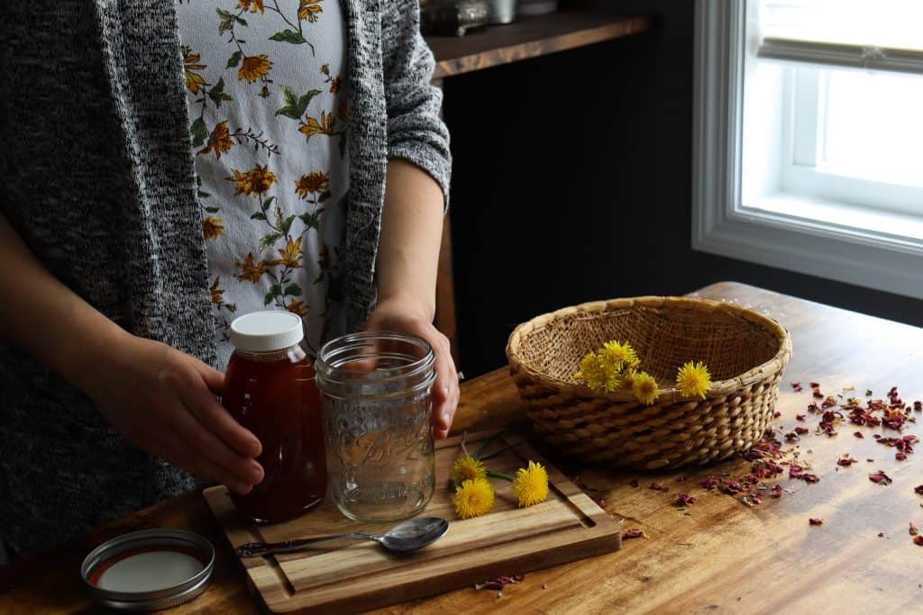 a woman prepared the ingredients and tools for dandelion infused honey. from left to right: honey, a spoon, a glass jar with a lid, and dandelions.