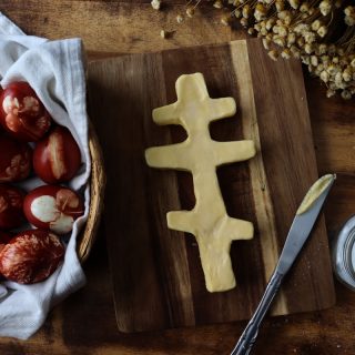 completed orthodox pascha butter cross on a wooden cutting board next to dried flowers, red eggs with floral prints on them, and salt in a glass jar. a butter knife rests nearby with butter on the edge of it!