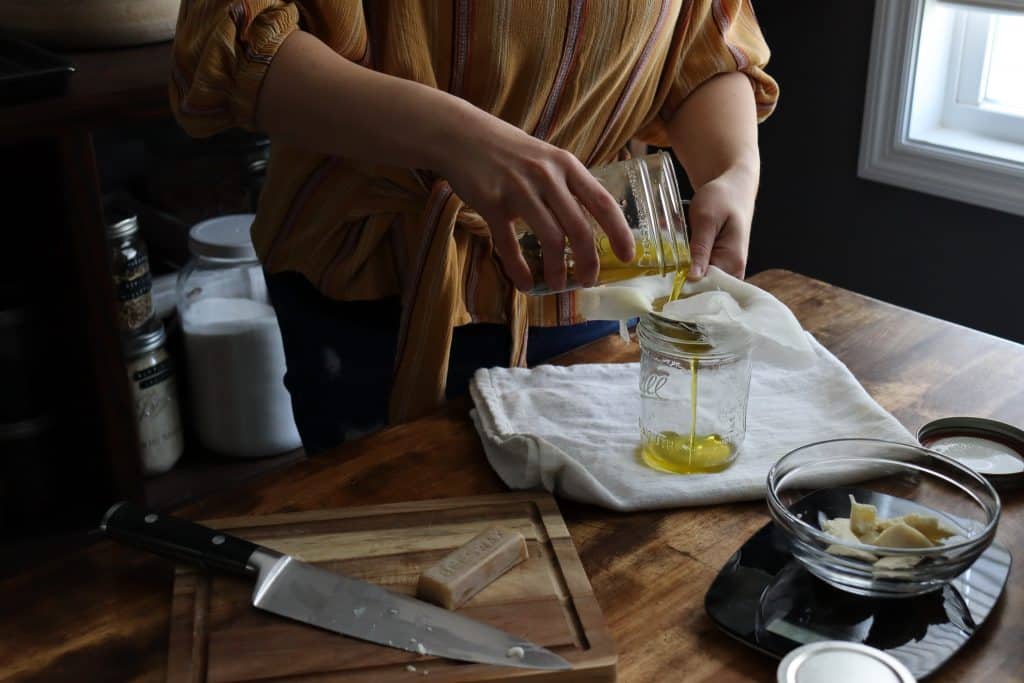 a woman is straining dandelion infused olive oil through a mesh strainer lined with cheese cloth
