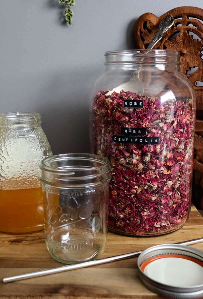 from right to left: raw honey, a glass jar, and dried rose petals In front lies a metal straw and a lid.