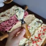 butter board with 5 butters on it. a woman holds a serving knife with some herb butter on it