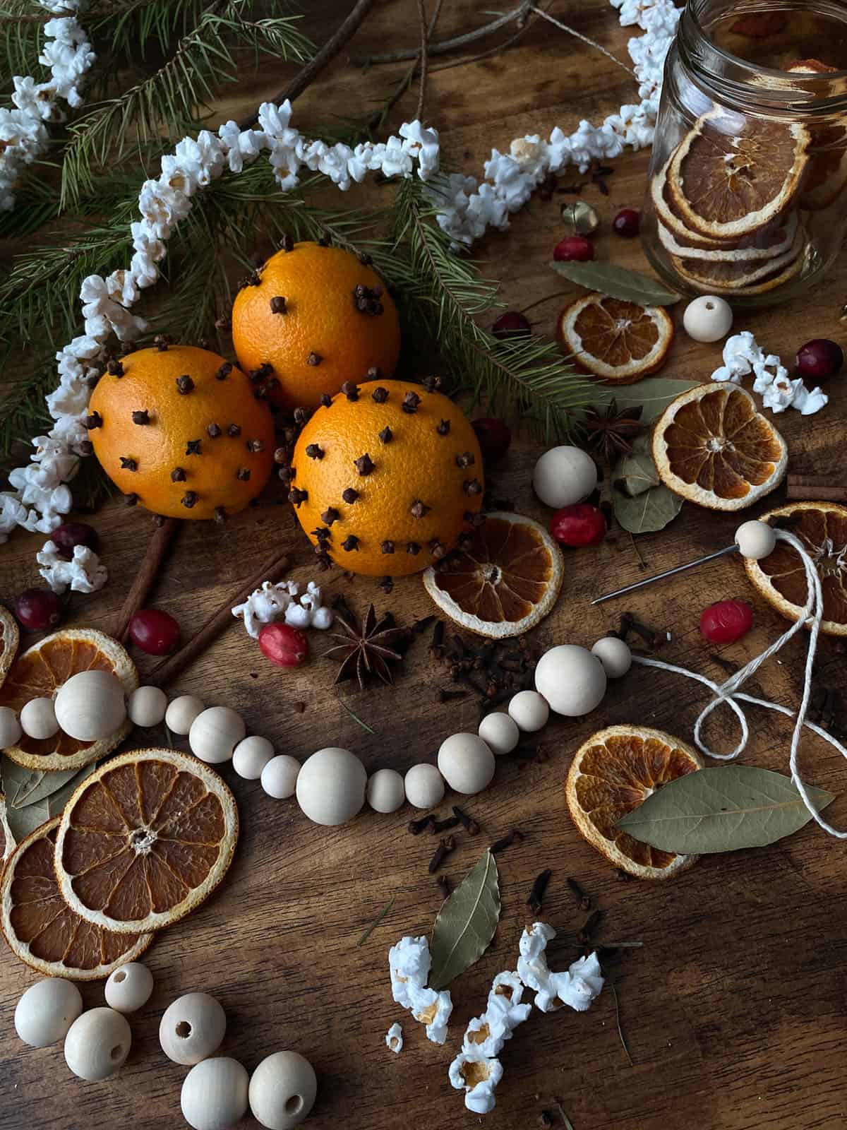 three orange pomanders are surrounded by a popcorn garland, pine branches, dried orange slices, cranberries, popcorn, star anise, and a string of wooden beads being made.