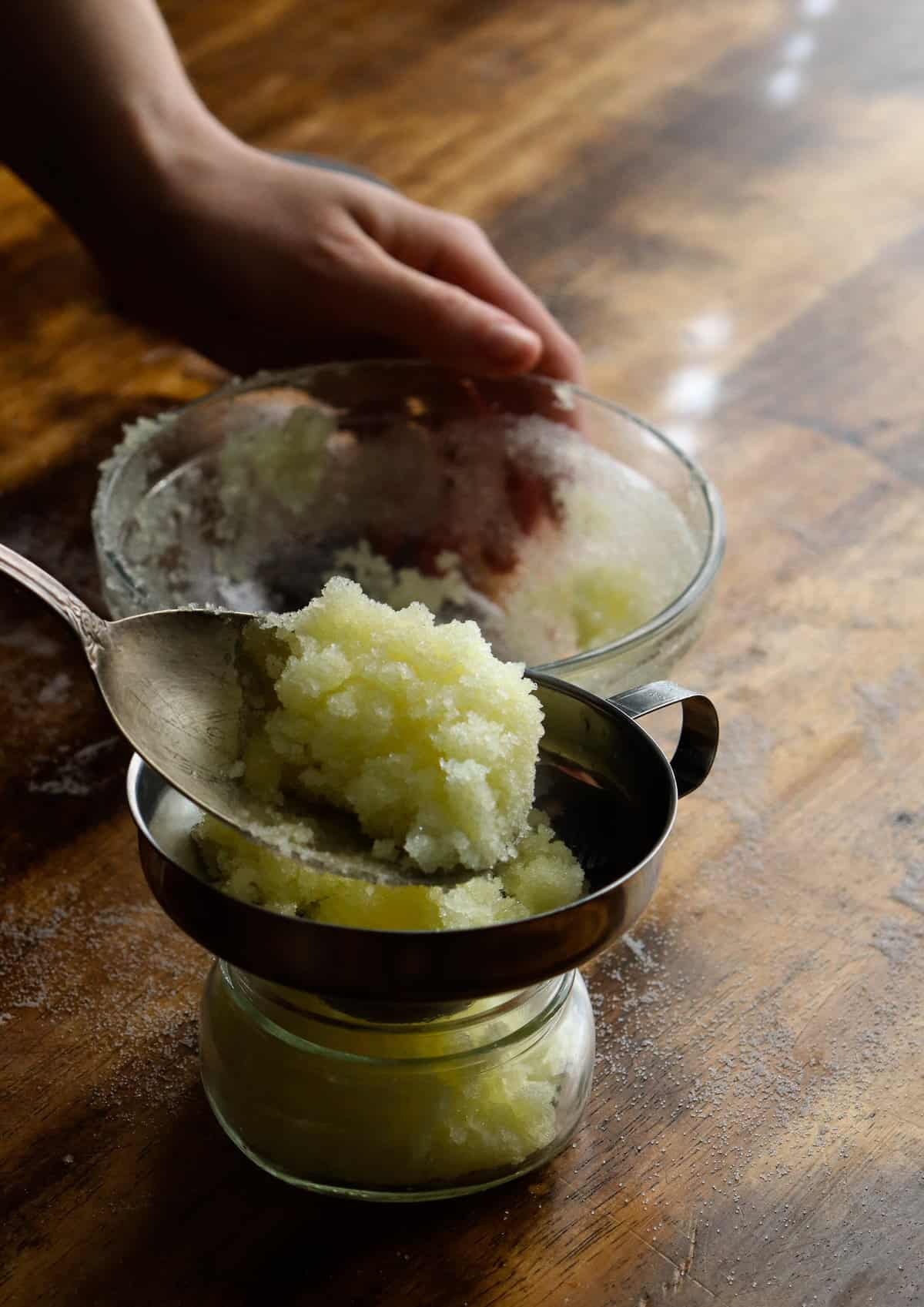 scooping salt scrub into a glass jar using a large silver spoon and a canning funnel