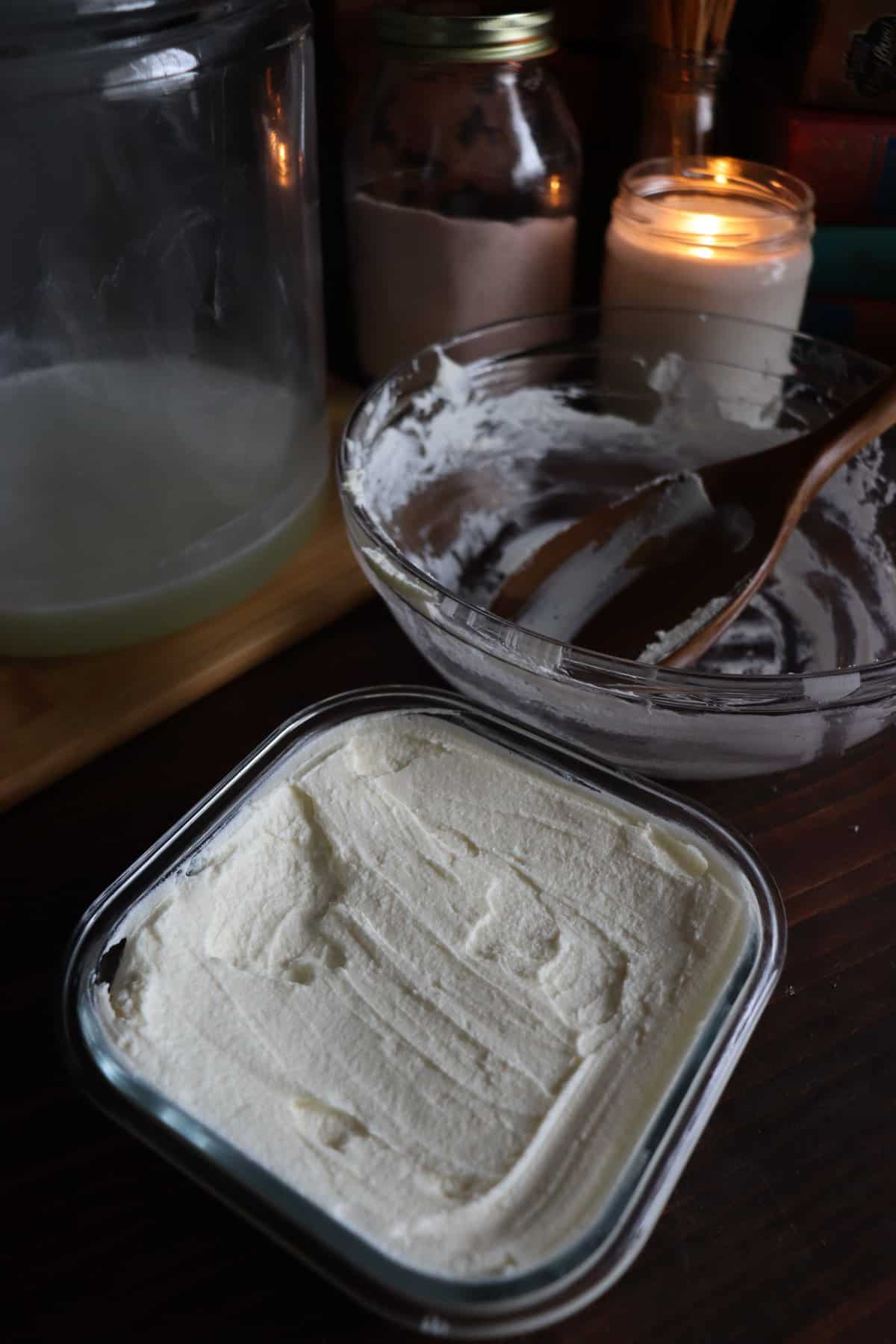 cream cheese in a glass container next to the bowl it was just in and a wooden spoon. the whey is still in the big glass jar it was dripping in. a candle is lit behind it all.