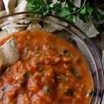 semi chunky salsa in a glass bowl. a chip is dipped in it. there is a cilantro plant above it and lots of tortilla chips!