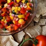a glass bowl of mango pico de gallo surrounded by chips and roma tomatoes