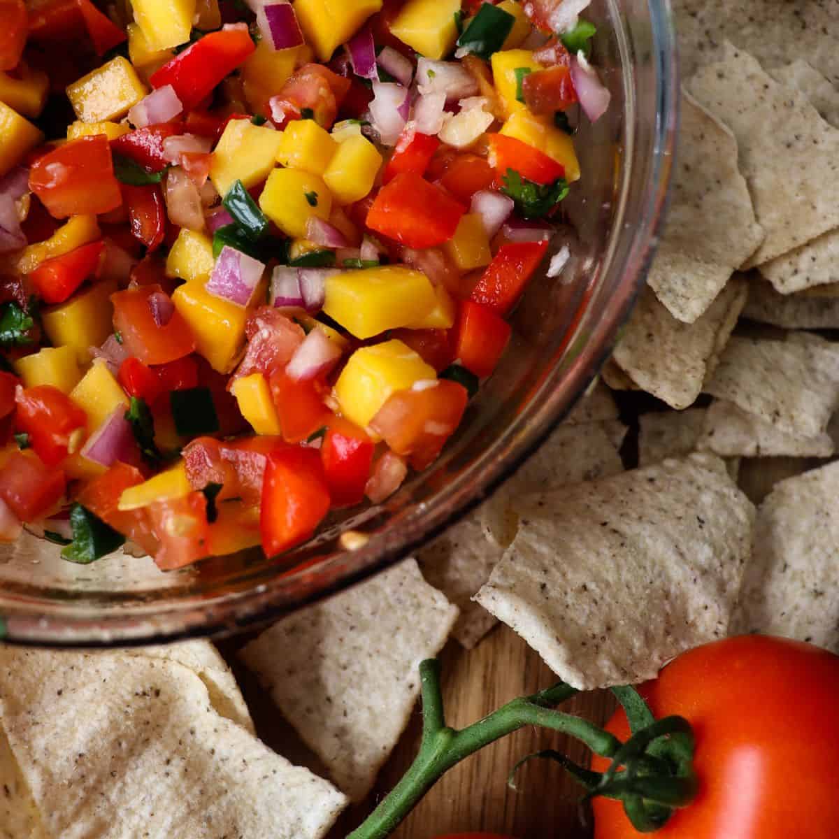 chunky mango pico de gallo in a glass bowl surrounded by chips and roma tomatoes