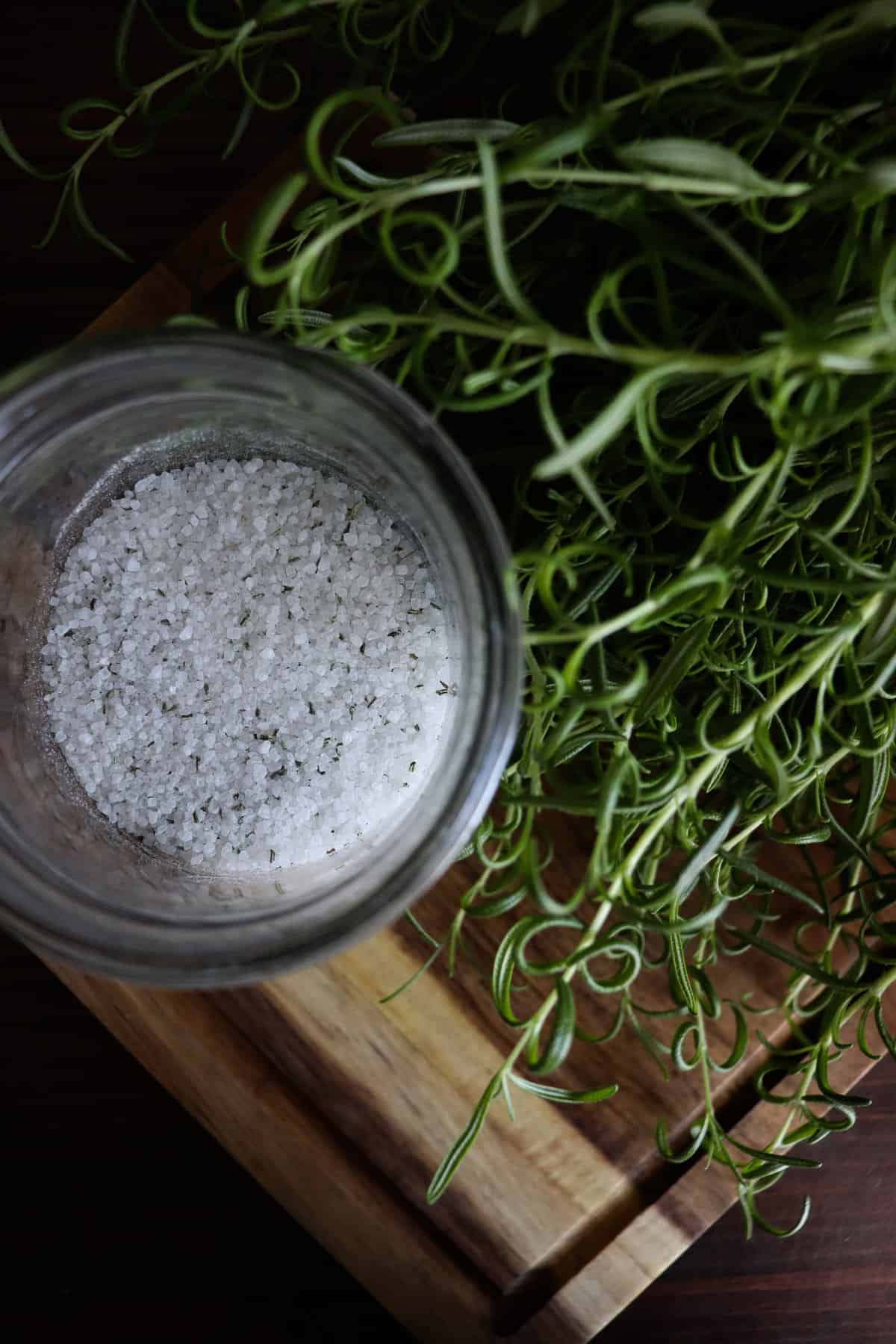 a thriving rosemary plant near some rosemary salt ready for a lid on a wooden cutting board
